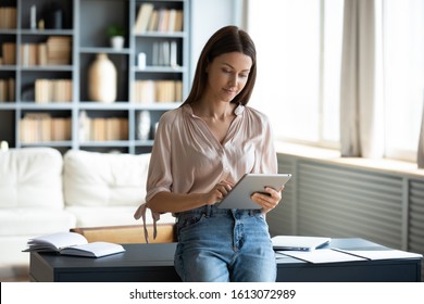 Smiling millennial woman stand using modern pad gadget at home browsing Internet or shopping, happy satisfied young female client surf web read news or chat on tablet device, technology concept