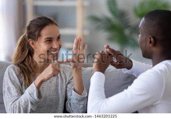 Smiling mixed ethnicity couple or interracial\
friends talking with sign finger hand language, happy two deaf and\
mute hearing impaired people communicating at home sit on sofa\
showing hand gestures