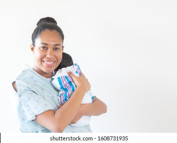 A Smiling Mixed Race African American Mother Wearing A Hospital Gown Holds Her Brand New Infant Baby On Her Shoulder Hugging And Cradling Him As He Sleeps In His Blanket Swaddle.
