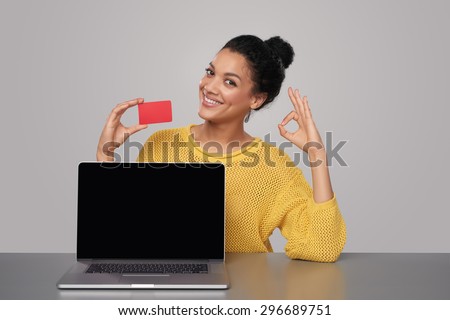 Smiling mixed race african american - caucasian woman showing blank black laptop computer screen and blank credit card, sitting at table and showing OK sign, over gray background