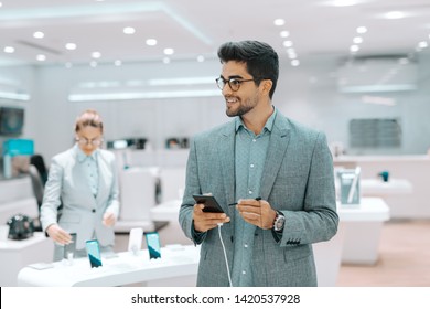 Smiling Mixed Race Bearded Man Dressed In Formal Wear Trying Out Smart Phone And Looking Away. Tech Store Interior.