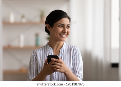 Smiling mixed race female stand with modern mobile phone in hand use app look aside think of pleasant message at social network. Content indian lady feel glad making awaited purchase online using cell