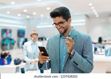 Smiling Mixed Race Man Trying Out New Smart Phone. Tech Store Interior, In Background Customer Standing Near Stand. Tech Store Interior.