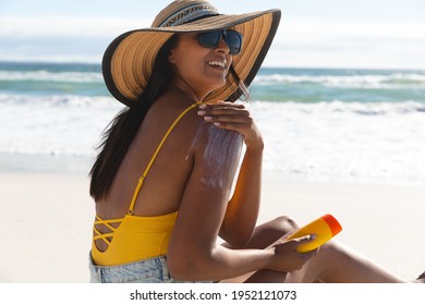 Smiling mixed race woman on beach holiday using sunscreen cream. healthy outdoor leisure time by the sea.