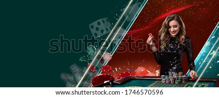 Smiling model in shiny black dress. Showing two red chips, posing at playing table on colorful background with flying money and dices. Poker, casino