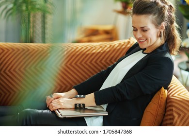 Smiling Modern 40 Years Old Woman In White Blouse And Black Jacket With Closed Laptop Sitting On Couch At Modern Home In Sunny Day.