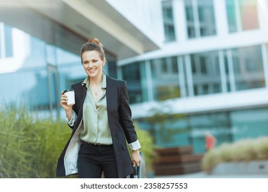 smiling modern 40 years old woman employee near office building in black jacket with cup of coffee walking. - Shutterstock ID 2358257553