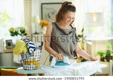 smiling modern 40 years old woman in silk blouse and beige pants with washed clothes basket at modern home in sunny day. ironing white shirt