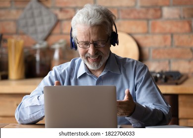 Smiling Modern Elderly 70s Man In Headphones Talk On Video Call On Laptop At Home. Happy Smart Mature Grandfather In Earphones Have Webcam Conversation Or Virtual Digital Conference On Computer.