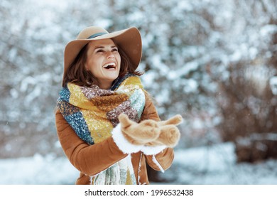 smiling modern female in brown hat and scarf with mittens in sheepskin coat catching snow outdoors in the city park in winter.