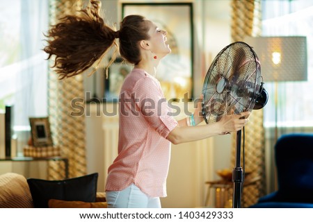smiling modern housewife in the modern living room in sunny hot summer day enjoying fresh air in the front of working fan.