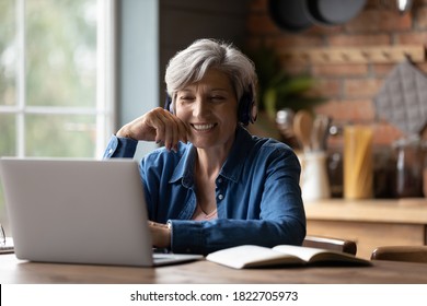 Smiling modern mature 60s Caucasian woman in earphones look at laptop screen watch webinar online at home. Happy senior grey-haired female have fun study on internet. Elderly technology concept.