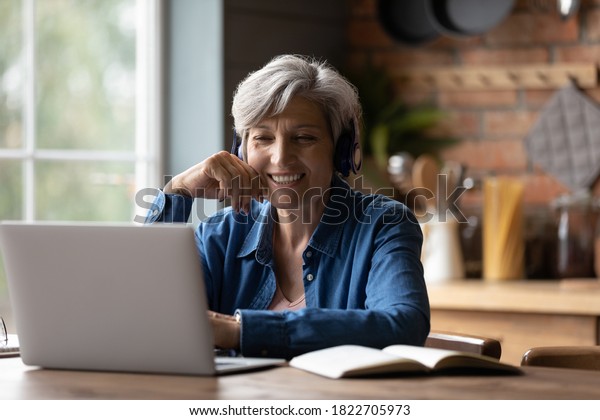 Smiling modern mature 60s Caucasian woman in\
earphones look at laptop screen watch webinar online at home. Happy\
senior grey-haired female have fun study on internet. Elderly\
technology concept.