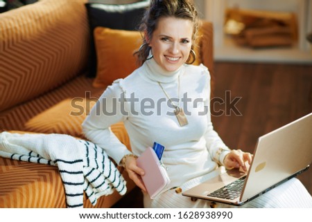 smiling modern middle age housewife in white sweater and skirt with credit card sitting near couch looking for new sweater at fashion online shop on a laptop in the modern house in sunny winter day.