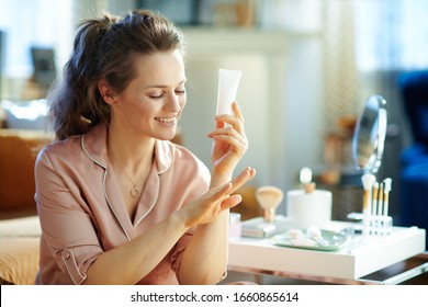 Smiling Modern Middle Age Woman In Pajamas With White Cosmetic Tube Applying Hand Cream Near Table With Toiletries At Modern Home In Sunny Winter Day.