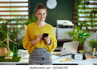smiling modern middle aged small business owner woman in yellow sweater in the modern green office using smartphone. - Shutterstock ID 2221041729