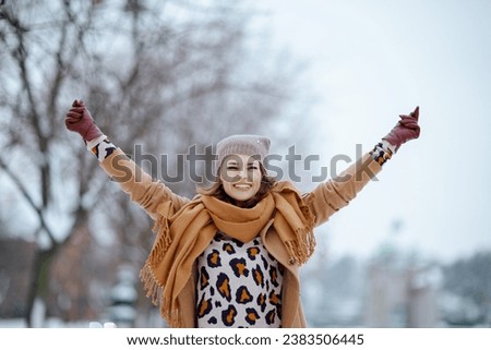 smiling modern middle aged woman in brown hat and scarf in camel coat with gloves rejoicing outside in the city in winter.