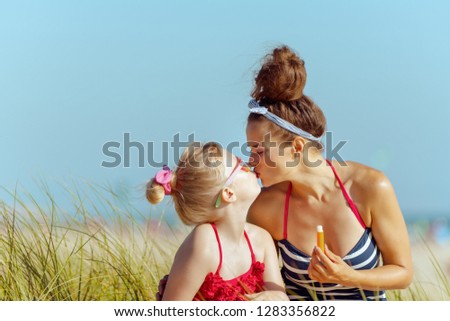 smiling modern mother and child in swimwear holding lipstick with spf and kissing on the ocean coast. lip balm with SPF sun protection to combat dry lips. getting vitamin D after long winter months.