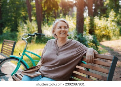 smiling modern old lady relaxing city park. Pensive senior gray haired woman casual sitting bench outdoors reading book. cycling forest park, bicycle, healthy active lifestyle after 50-60 years - Powered by Shutterstock