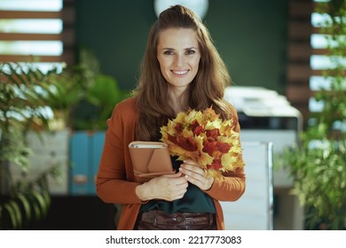 Smiling Modern Small Business Owner Woman With Notebook And Autumn Yellow Leaves In The Modern Green Office.