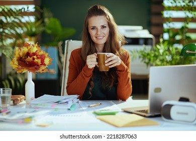 Smiling Modern Small Business Owner Woman With Autumn Yellow Leaves And Cup Of Hot Cocoa In The Modern Green Office.