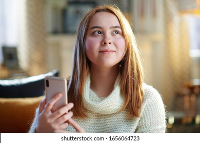 Smiling Modern Teenage Girl With Red Hair In White Sweater At Modern Home In Sunny Winter Day Checking Social Media Account On A Smartphone.