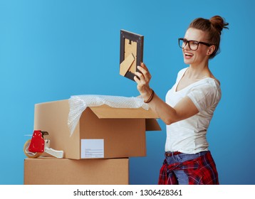 Smiling Modern Woman In White T-shirt Near Cardboard Box Looking At The Photo Frame Against Blue Background