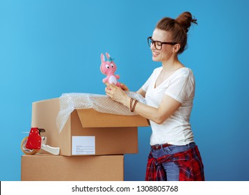 Smiling Modern Woman In White T-shirt Near Cardboard Box Looking At Old Toy Against Blue Background