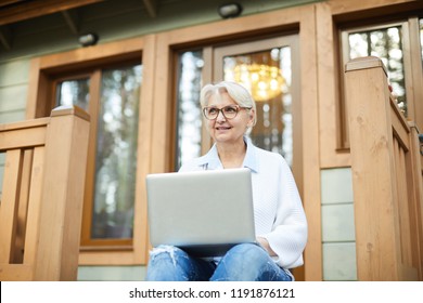 Smiling modern working senior lady in glasses sitting on porch of cottage and looking into distance while working on project using laptop - Powered by Shutterstock