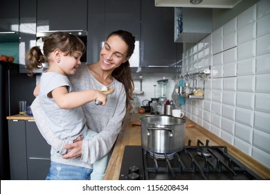 Smiling mom holding cute kid girl hepling mum in the kitchen, happy family of young single mother and daughter talking having fun cooking together, happy mommy teaching child preparing food