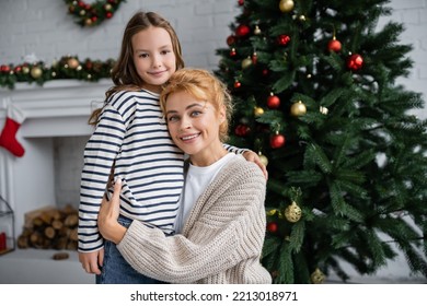 Smiling Mom In Knitted Cardigan Hugging Daughter Near Christmas Tree At Home