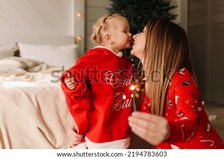 Smiling mom and little daughter in red pajamas hug, kiss and burn a sparkler celebrating the Christmas holiday in a cozy bedroom with a Christmas tree at indoor house. Selective focus