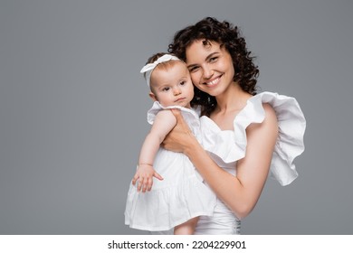 Smiling Mom Looking At Camera While Holding Baby Girl In White Clothes Isolated On Grey