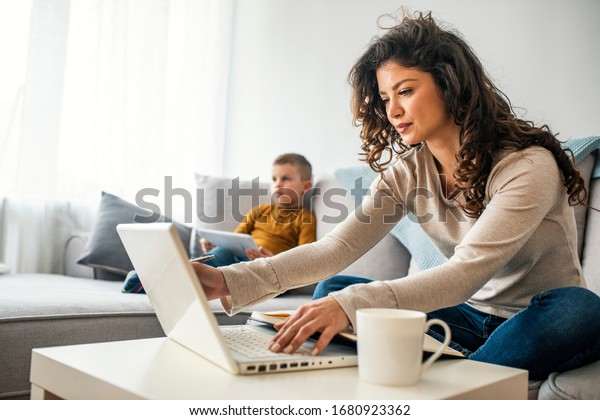 Smiling mom working at
home with her child on the sofa while writing an email. Young woman
working from home, while in quarantine isolation during the
Covid-19 health crisis