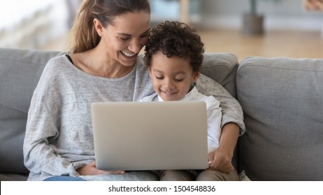 Smiling mother with African American son using laptop close up, happy mum and preschool child sitting on sofa at home, watching cartoons online or play video game, family having fun