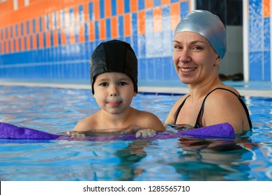 Smiling Mother In Black Bathing Suit And Silver Rubber Hat Teach To Swim Happy Little Girl With Pool Noodle In Public Indoor Swimming Pool. Family Holiday Concept, Training Of Champion