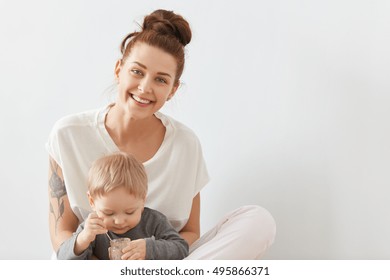 Smiling Mother Caring About Her Three-years Old Child On White Background. Lovely Blond Kid In Grey Shirt Eating Childhood Nutrition From Glass Can With Little Spoon. His Mom Looking At Camera.