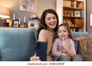 Smiling mother and daughter on video call using digital phone at home. while making video call and waving hello sitting on sofa. Woman and her cute little kid doing video conference with grandparents. - Powered by Shutterstock