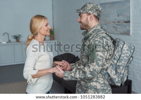 smiling mother and grown son in military uniform holding hands together at home