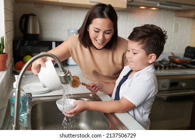 Smiling Mother Helping Son To Rinse Dishes Under Tap Water After Cleaning