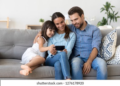 Smiling Mother Holding Smartphone Taking Family Selfie, Watching Video, Making Online Call Looking At Phone Screen, Shopping In Mobile App, Recording Vlog Embracing Child Daughter And Husband At Home