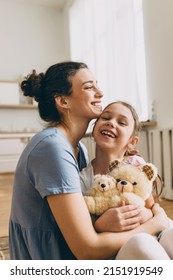 Smiling mother hugging cute little girl holding teddy bear toy, both laughing while cuddling, girl sitting on mom's knees, female tickling her child. Human relationships. Carefree childhood