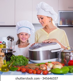 Smiling Mother And Little Daughter In Chefs Hat With Crockpot In Home Kitchen
