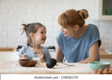 Smiling mother looking at little daughter and feeding her after bake cookies standing at table in kitchen. Happy caring mum and adorable girl eating flour and drink milk, preparing dinner.
