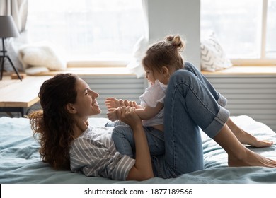 Smiling mother lying in cozy bed play and talk with cute little toddler daughter. Happy young Caucasian mom relax in home bedroom feel playful engaged in funny activity together with small girl child.