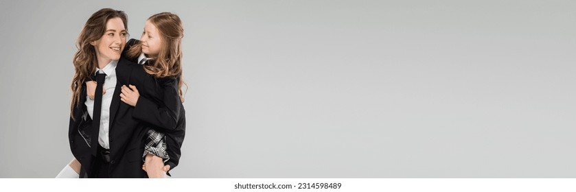 smiling mother piggybacking her daughter, working mom in business attire and schoolgirl in uniform on grey background in studio, modern parenting, fashionable family, having fun, banner - Powered by Shutterstock