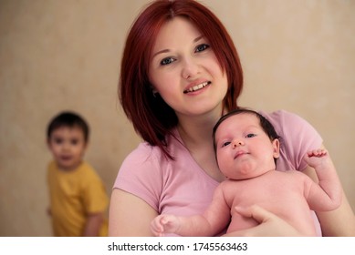 Smiling Mother Posing With Infant Child. Elder Toddler Kid Is Standing Behind, Blurred And Forgotten. Parenting Issues When New Baby Arrived, Young Mom Time Management, Family Relationships Concept