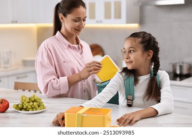 Smiling mother putting lunch box into daughter`s backpack in kitchen. Preparing for school - Powered by Shutterstock