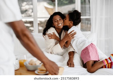 Smiling mother receiving a kiss from her daughter on a cozy morning, dad serving breakfast in bed, bright room. - Powered by Shutterstock