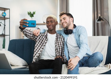 Smiling Multi Ethnic Couple Of Two Guys Sitting Together On Couch And Using Modern Smartphone For Taking Selfie. Fun, People And Gadgets Concept.